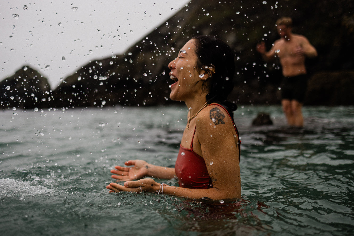 Woman in red swimming costume, stood in chest-high sea-water, invigoratingly throws up fresh ocean spray.