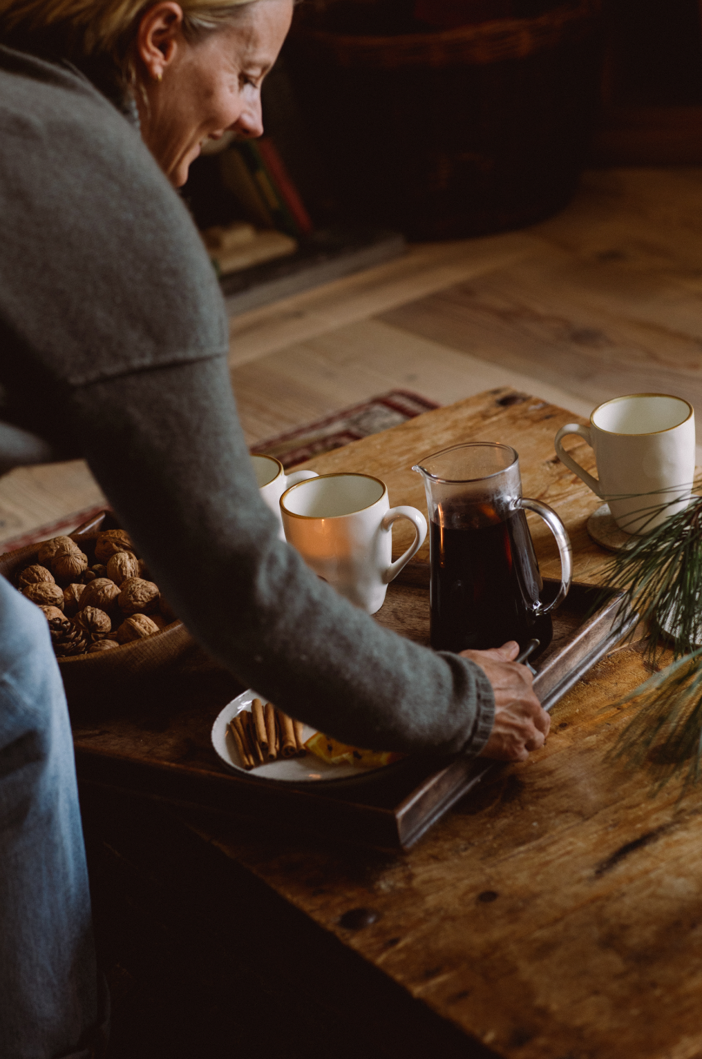Tray with jug of mulled wine, mugs, plate of cinnamon sticks and bowl of walnuts being placed on wooden table in cosy festive lounge.