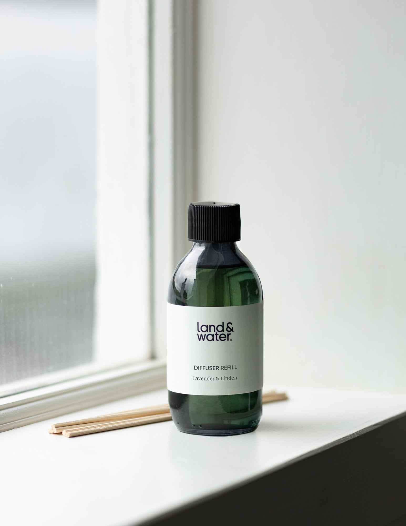 A 200ml bottle of lavender and linden essential oil diffuser refill by Land & Water, placed on a windowsill with reeds next to it.