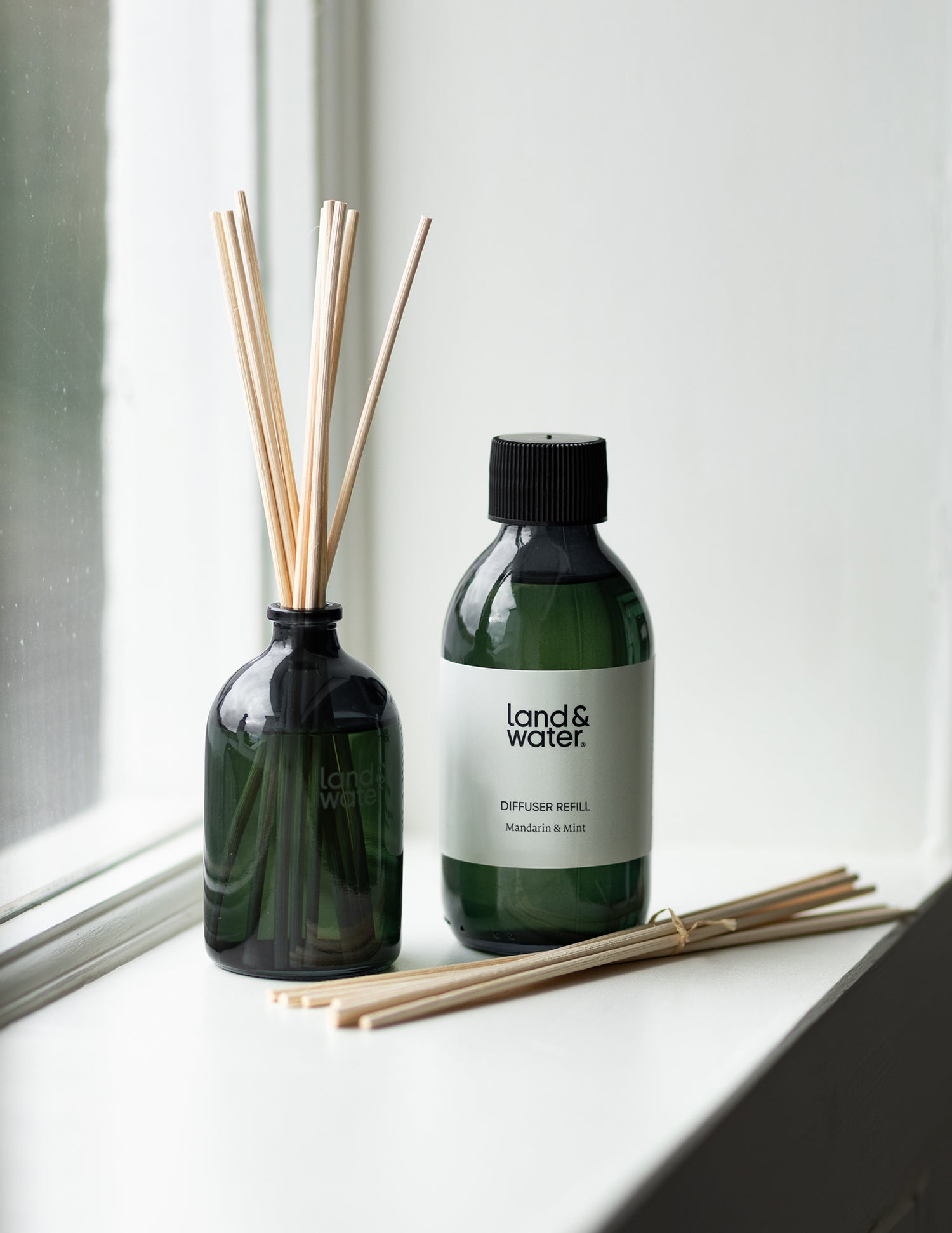 A land&water reed diffuser with a refill bottle and reed sticks lying on the window sill.