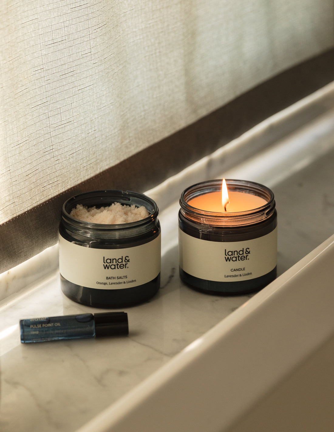 A set of bath and body products including bath salts, a pulse point oil, and a lit candle providing a warm glow. The products are placed on a marble surface.