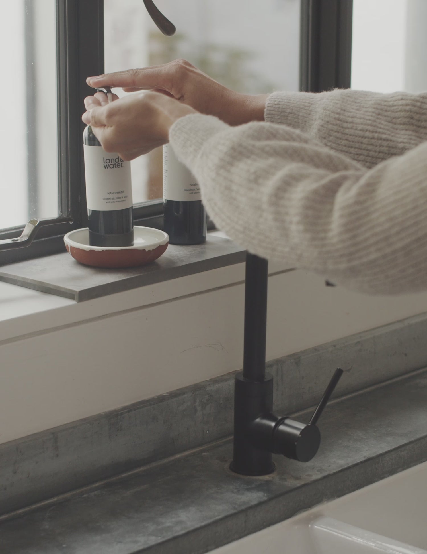 A short film clip of a woman wearing a soft oatmeal jumper, leaning over a kitchen sink washing her hands with land&water hand wash.