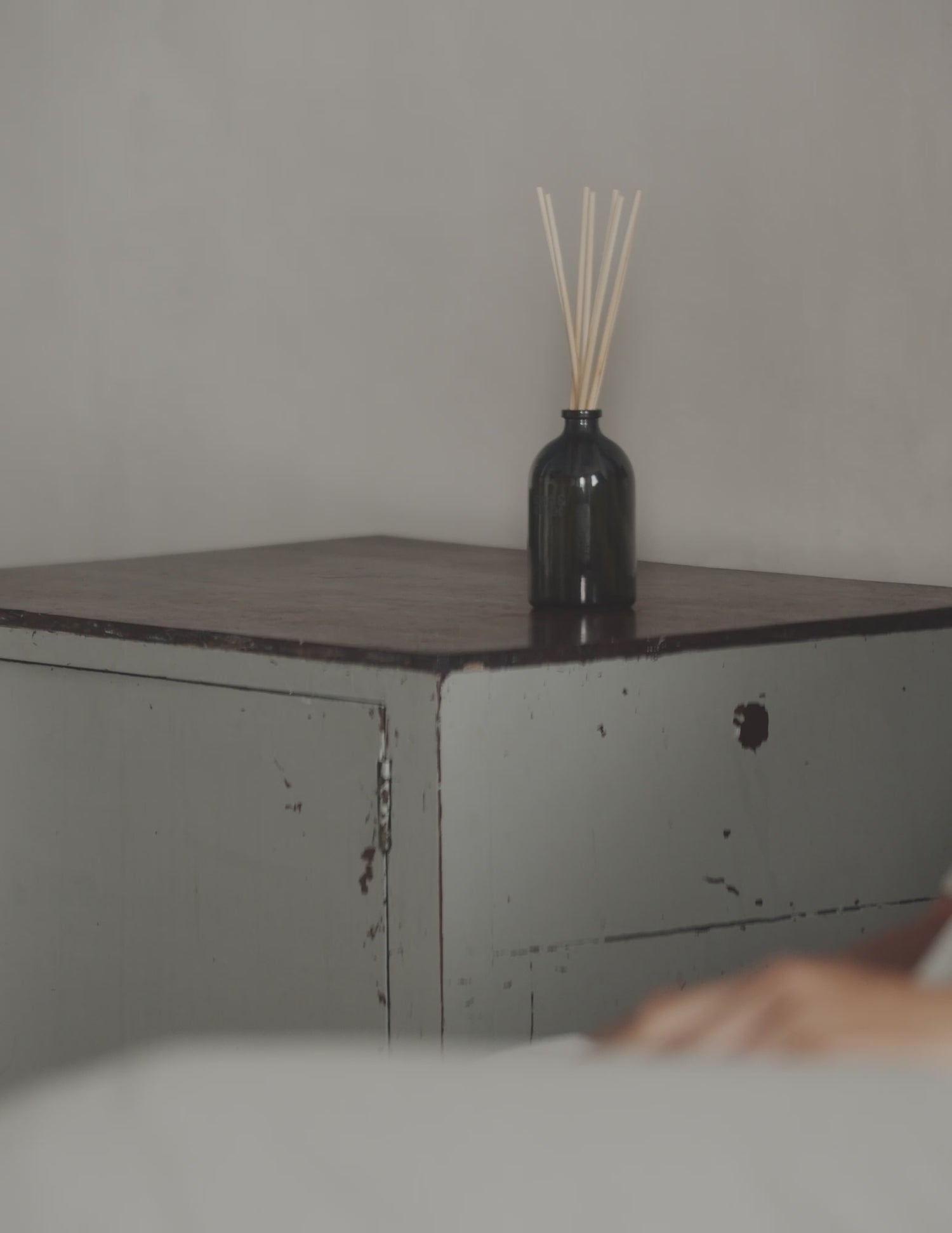 A landwater reed diffuser on a bedside table, with a woman walking past from the bed in pyjamas.