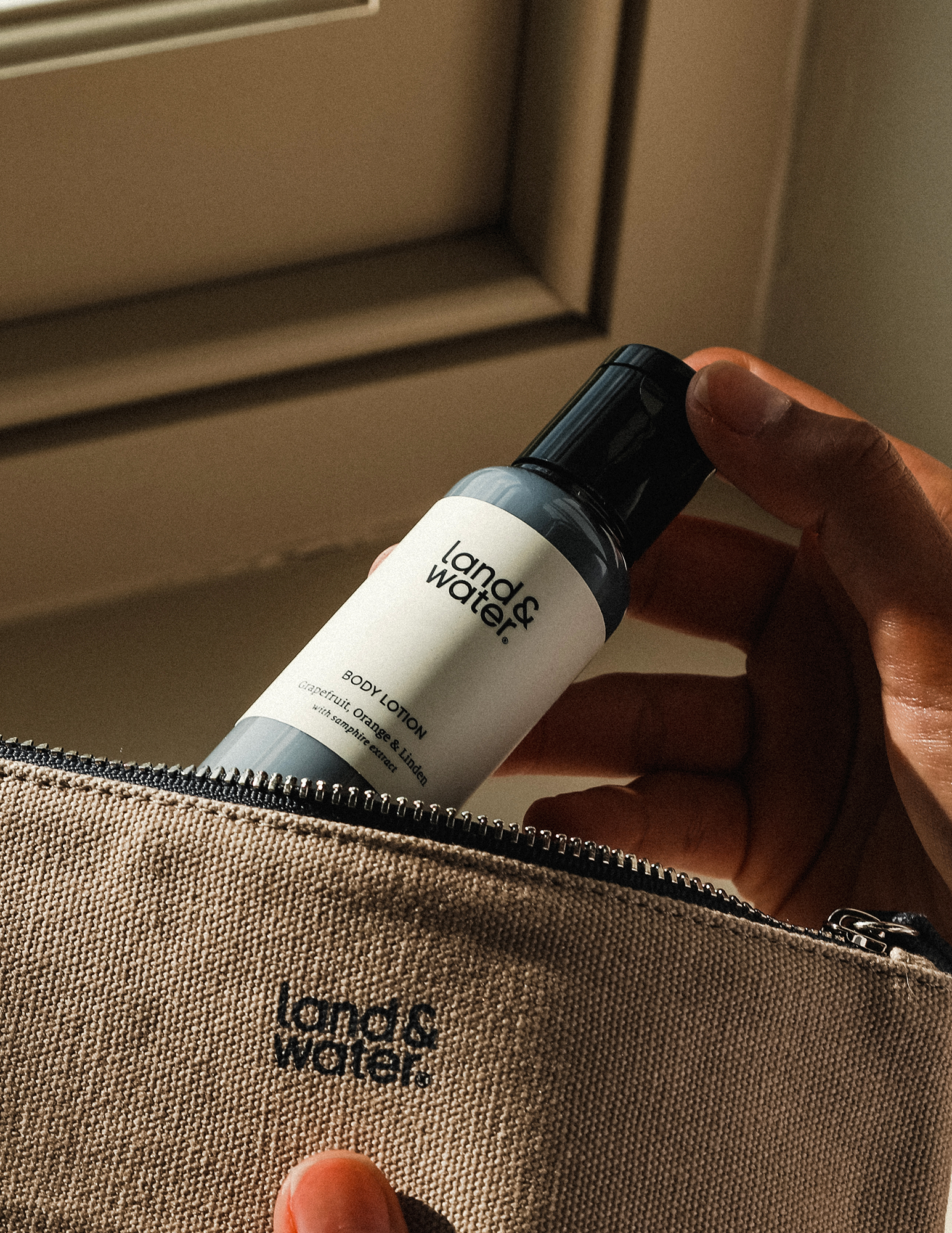 A hand holding a travel-sized body lotion bottle with the cap on, in front of a zippered pouch with the brand &