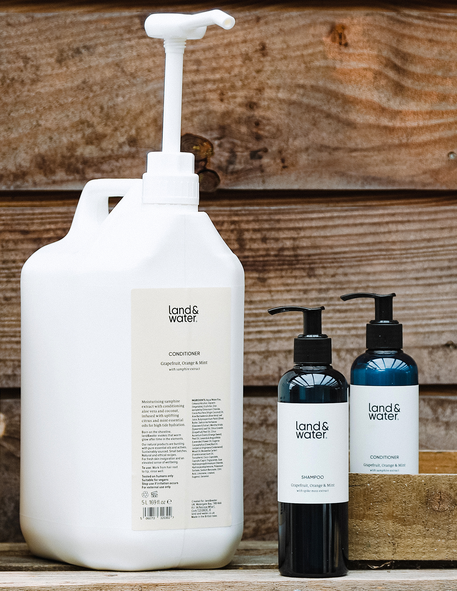 A 5L white bottle of conditioner with a pump dispenser, accompanied by smaller bottles of shampoo and conditioner with pumps on a wooden surface.