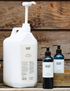 A 5L white bottle of conditioner with a pump dispenser, accompanied by smaller bottles of shampoo and conditioner with pumps on a wooden surface.