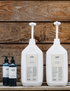 Two large white bottles with dispensers containing body lotion and body wash, alongside smaller bottles. Set against a wooden background.