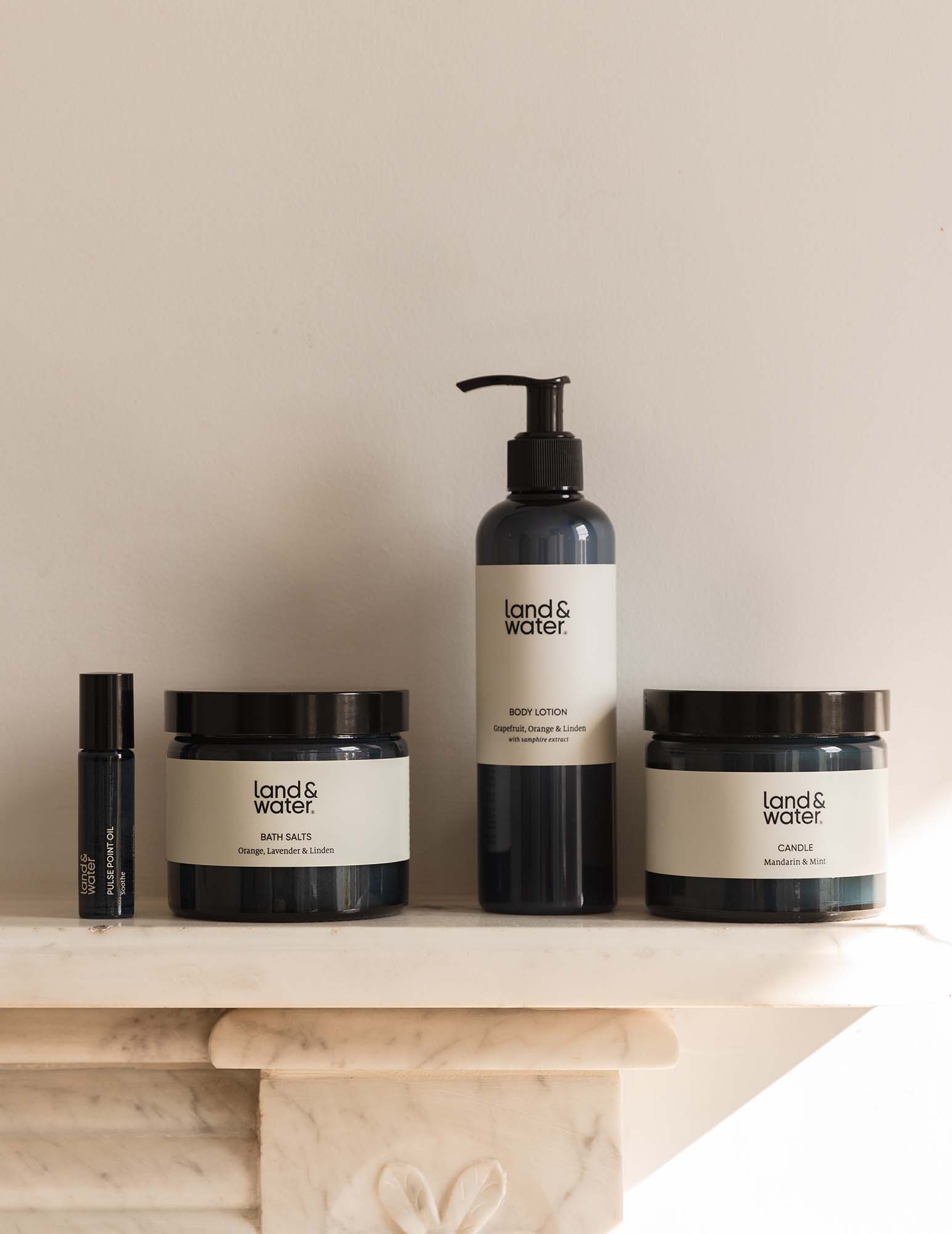 A collection of natural bath and body products including a candle, bath salts, pulse point oil, and body lotion, arranged on a marble surface.