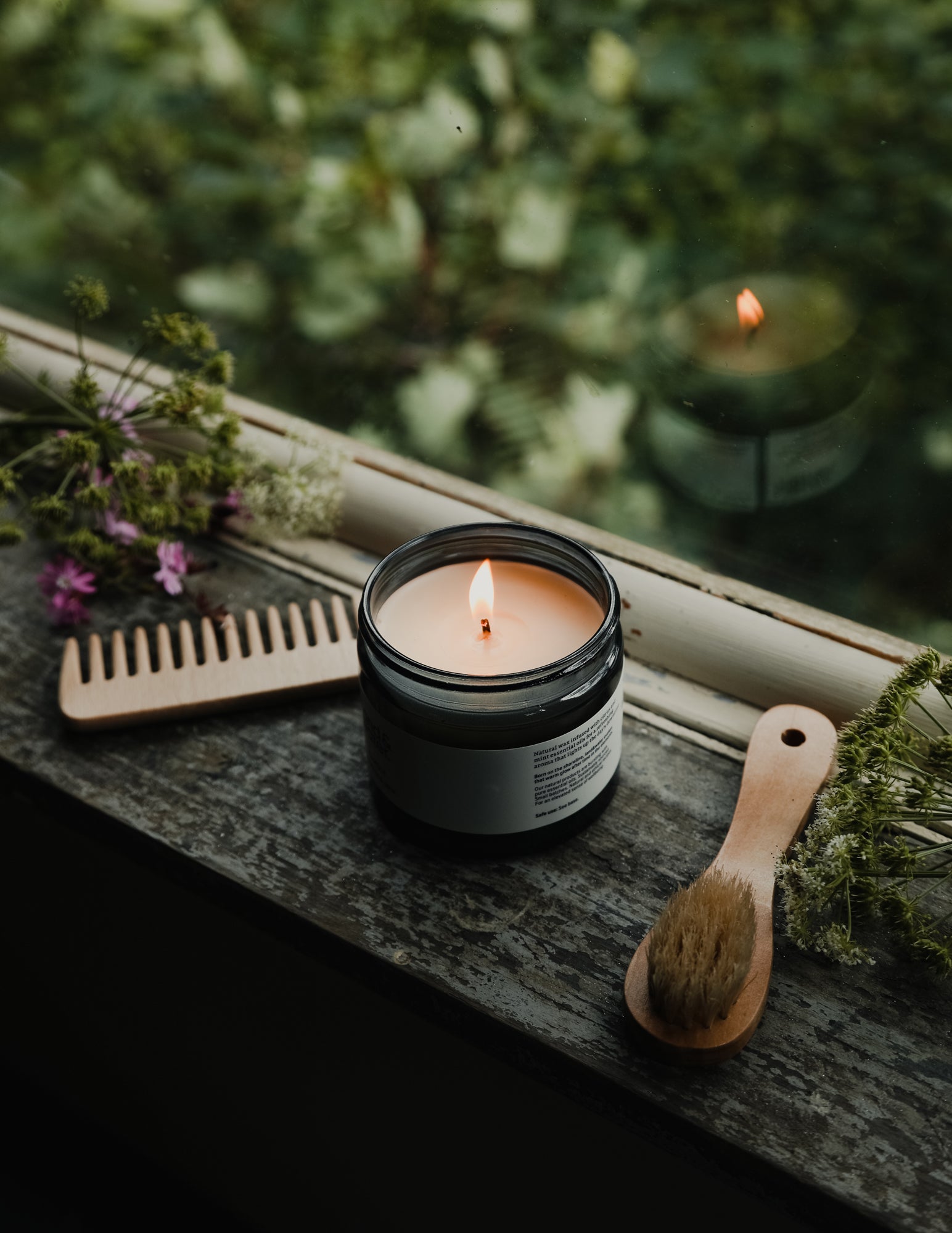 A lit land&water candle on a rustic wooden windowsill with flowers, wooden comb and brush with greenery through the window.