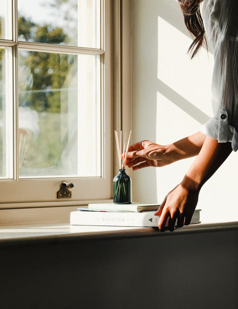 A woman adjusting the reeds in a reed diffuser on a sunny window sill.