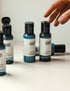 A collection of travel-sized bottles of hand sanitizer, body wash, body lotion, shampoo, and conditioner, arranged on a white surface with a hand reaching for one of the bottles.