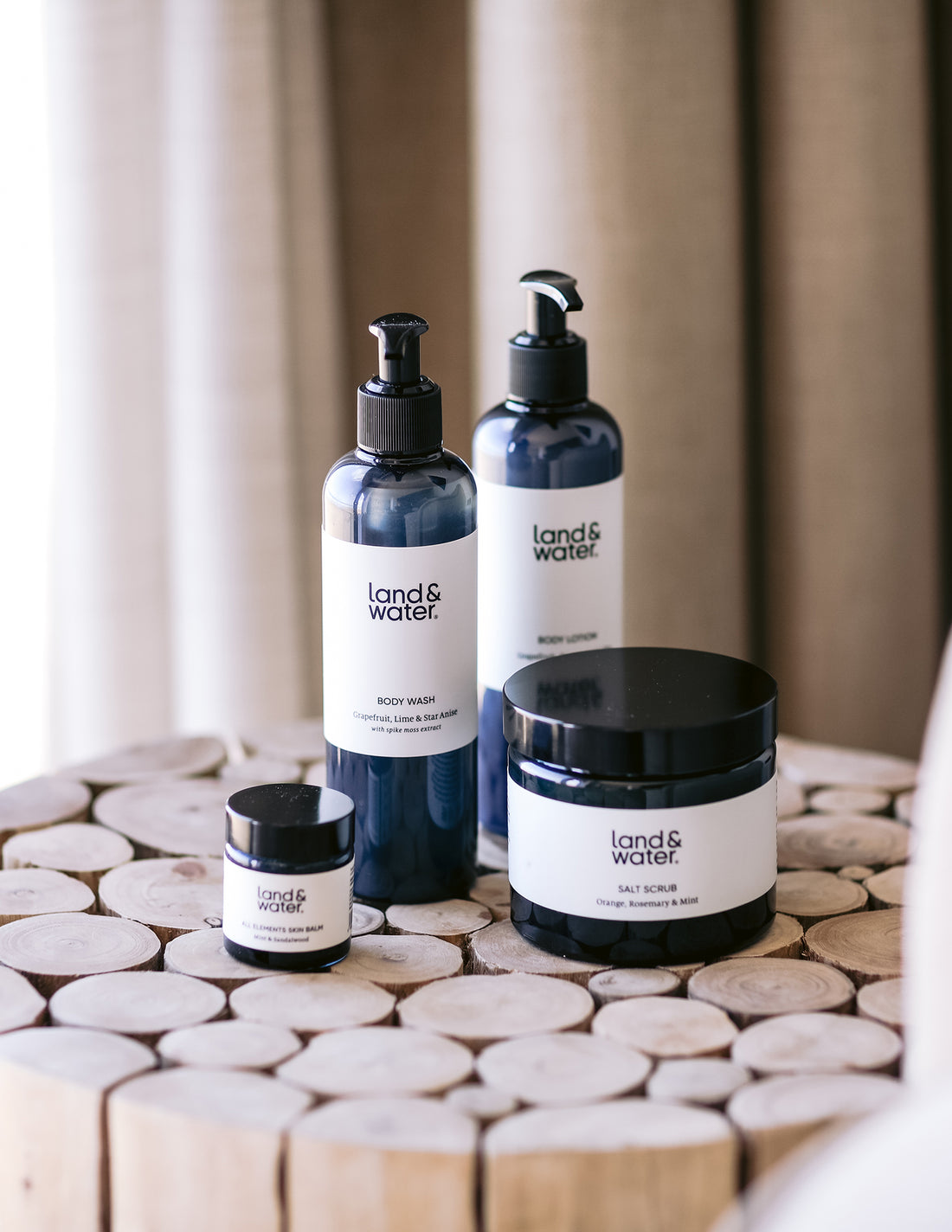 A collection of bath and body products from land&water, including a body wash, body lotion, salt scrub, and skin balm, displayed on a wooden surface with a window in the background.