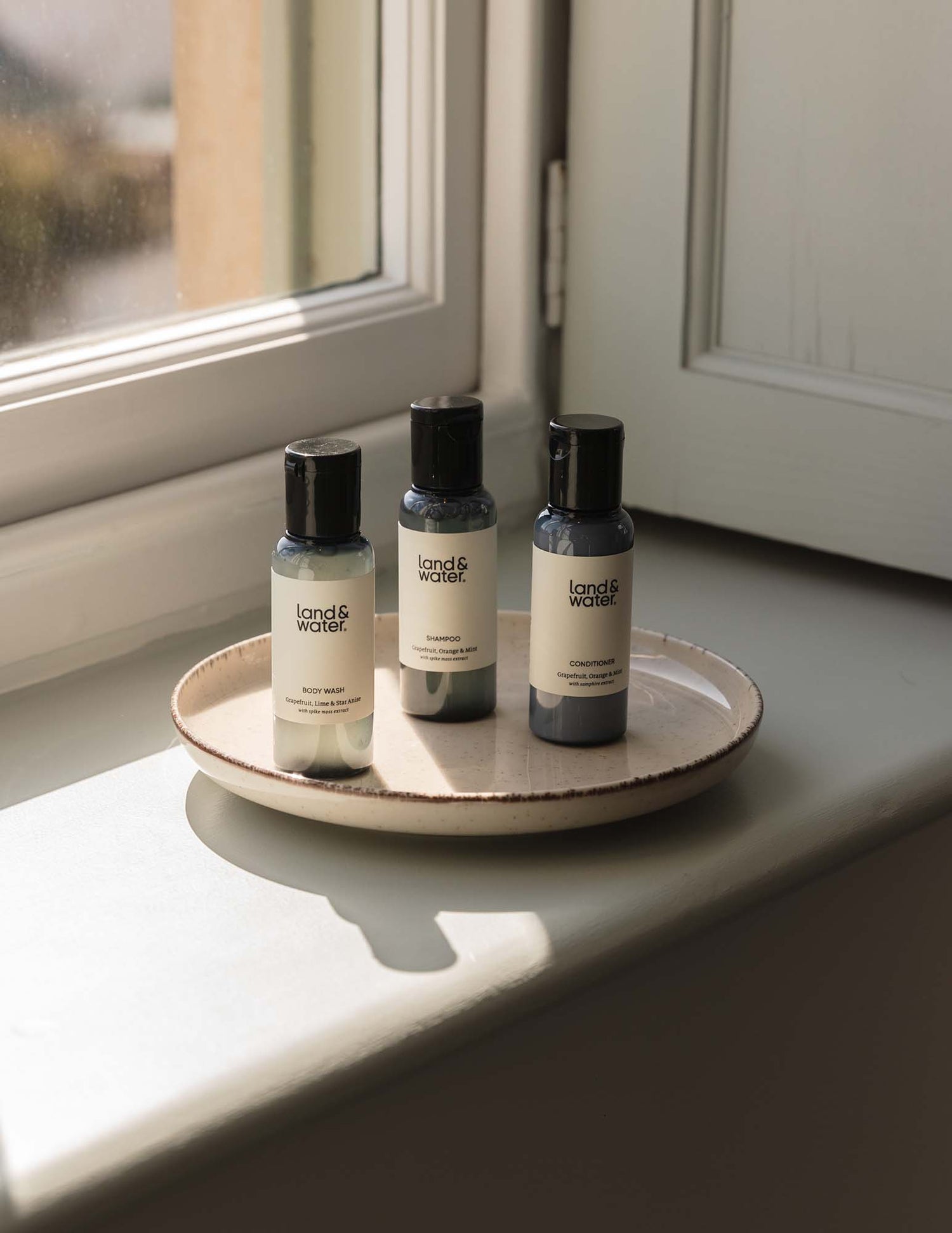 Three travel-sized bottles of body wash, shampoo and conditioner placed on a ceramic plate on a light windowsill.