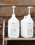 Two large white plastic bottles with dispensers containing shampoo and conditioner, placed on a wooden shelf against a wooden background.