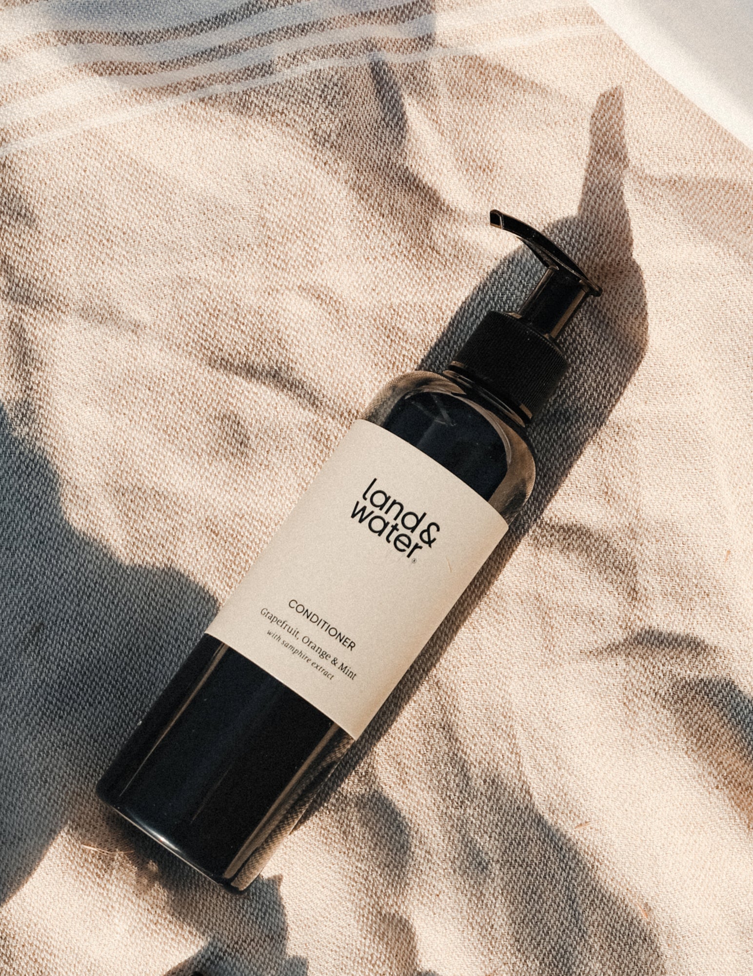 A bottle of land& water Conditioner 250ml, lying on a textured towelling surface in the sunlight.