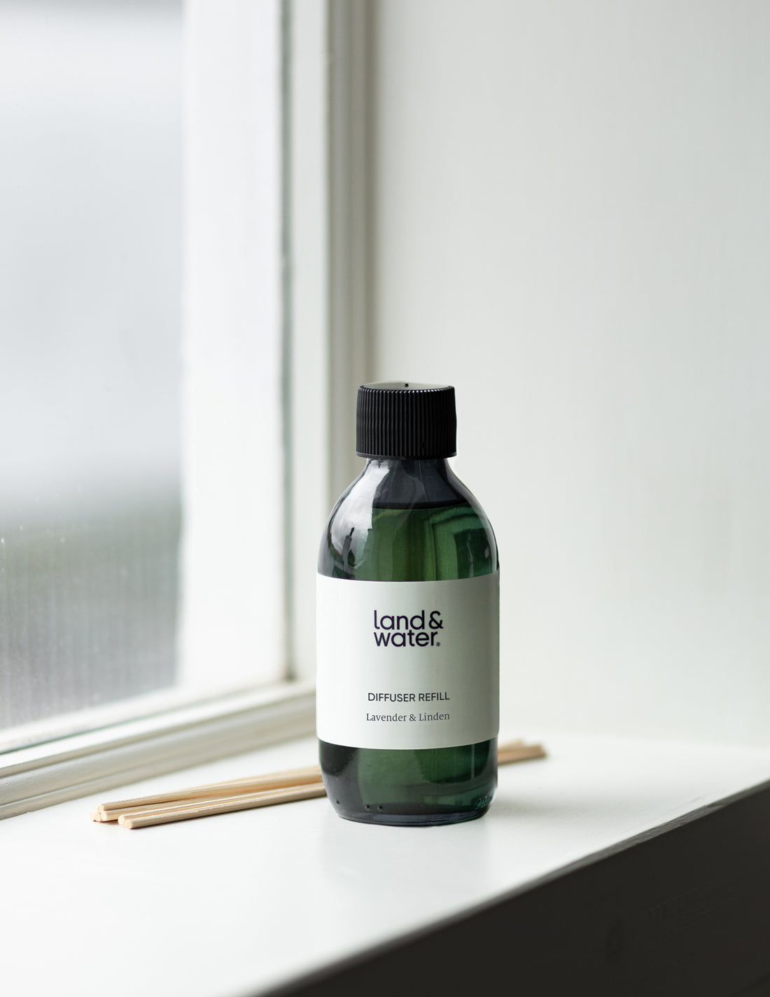 A 200ml bottle of lavender and linden essential oil diffuser refill by Land & Water, placed on a windowsill with reeds next to it.