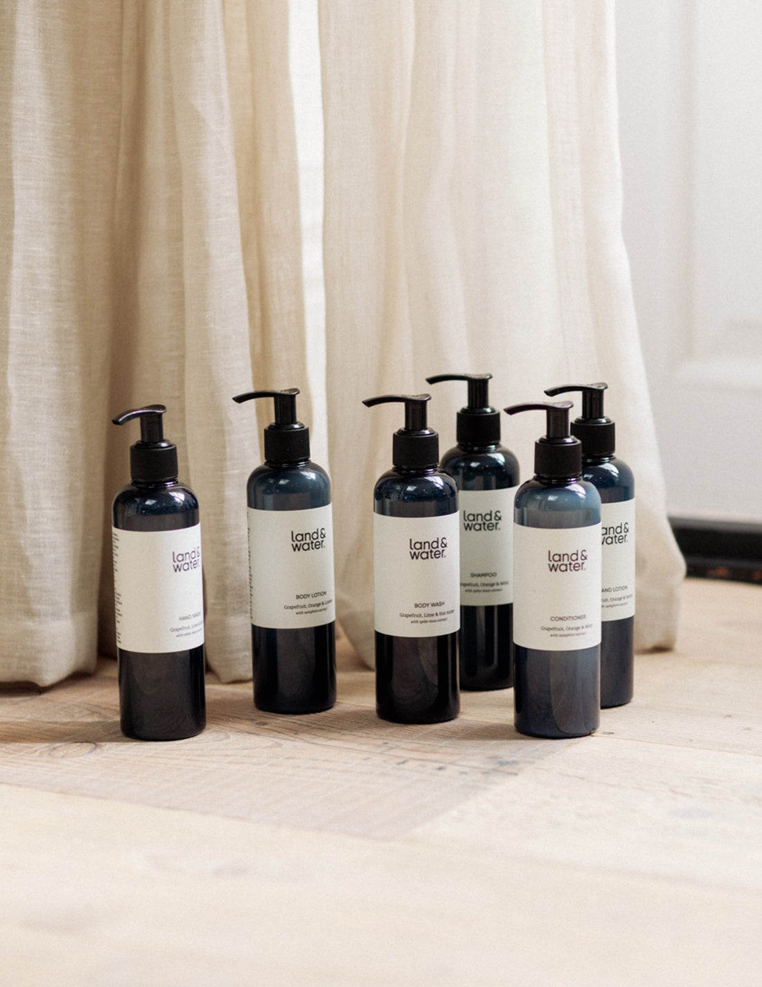 A series of blue pump bottles with cream labels, arranged in a line on a light-colored floor; hand wash, hand lotion, body wash, body lotion, shampoo, conditioner.