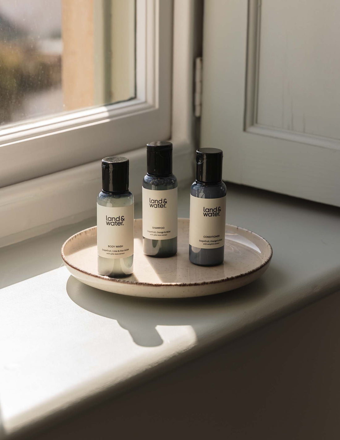 Three travel-sized bottles of body wash, shampoo and conditioner placed on a ceramic plate on a light windowsill.
