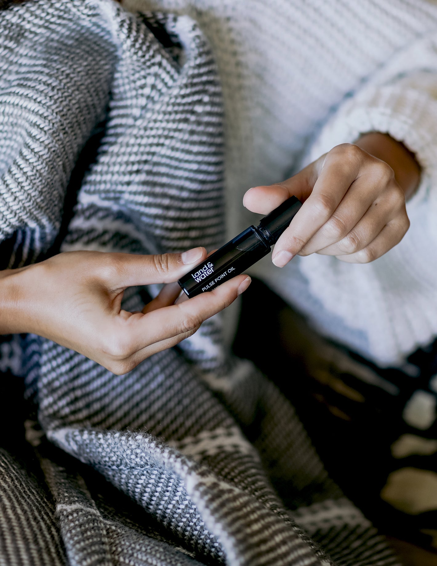 A woman's hands holding a pulse point oil, taking off the lid. There's a cosy blanket on her lap. 
